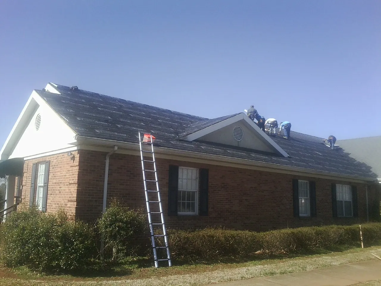 Crew tearing shingles off a brick ranch roof with a ladder set — Metal Roof Repair work by our crew in Pittsford