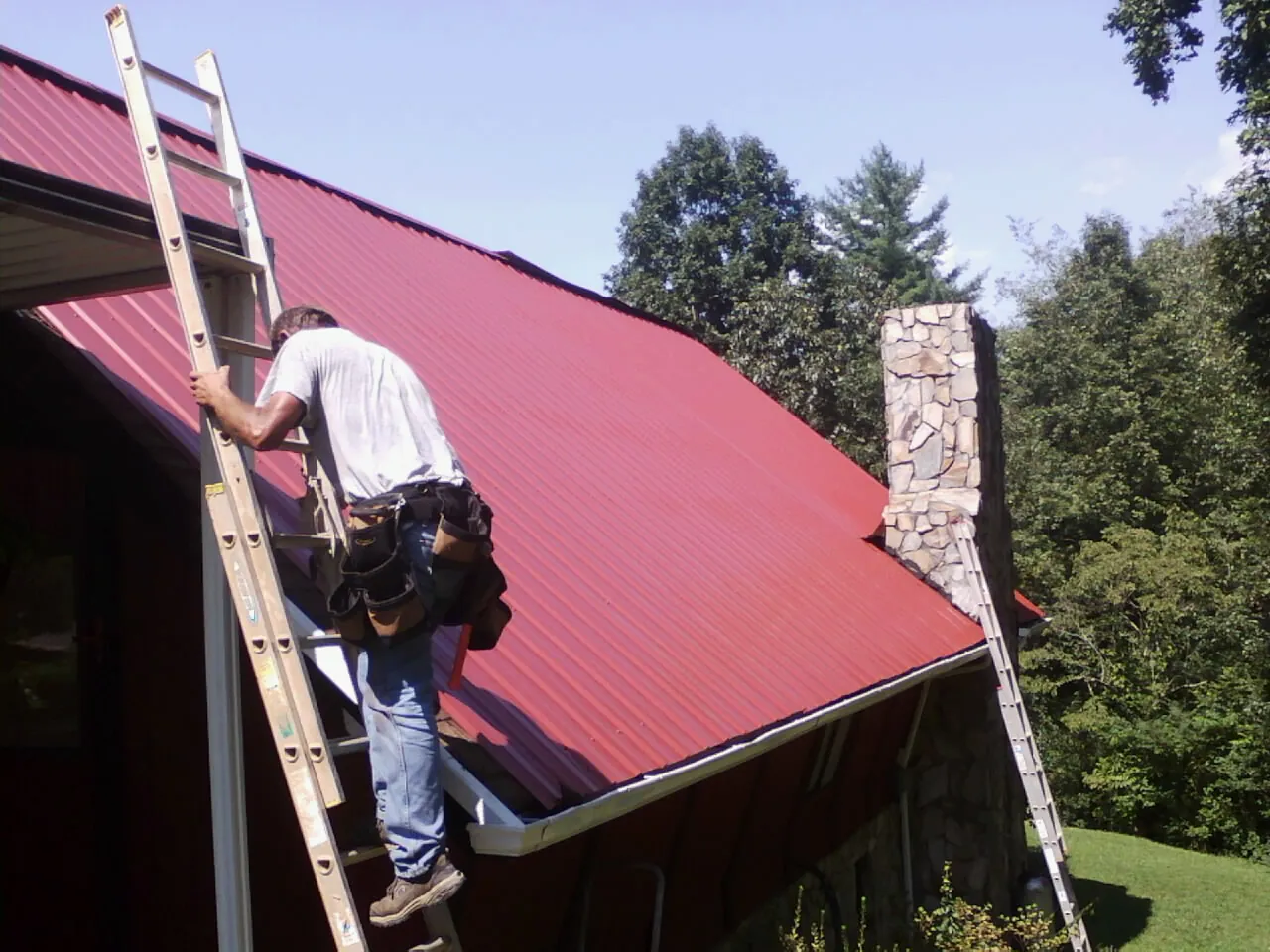 Roofer on a ladder beside a red metal roof and stone chimney — Soffit & Fascia Repair by Roofing Brothers in Pittsford